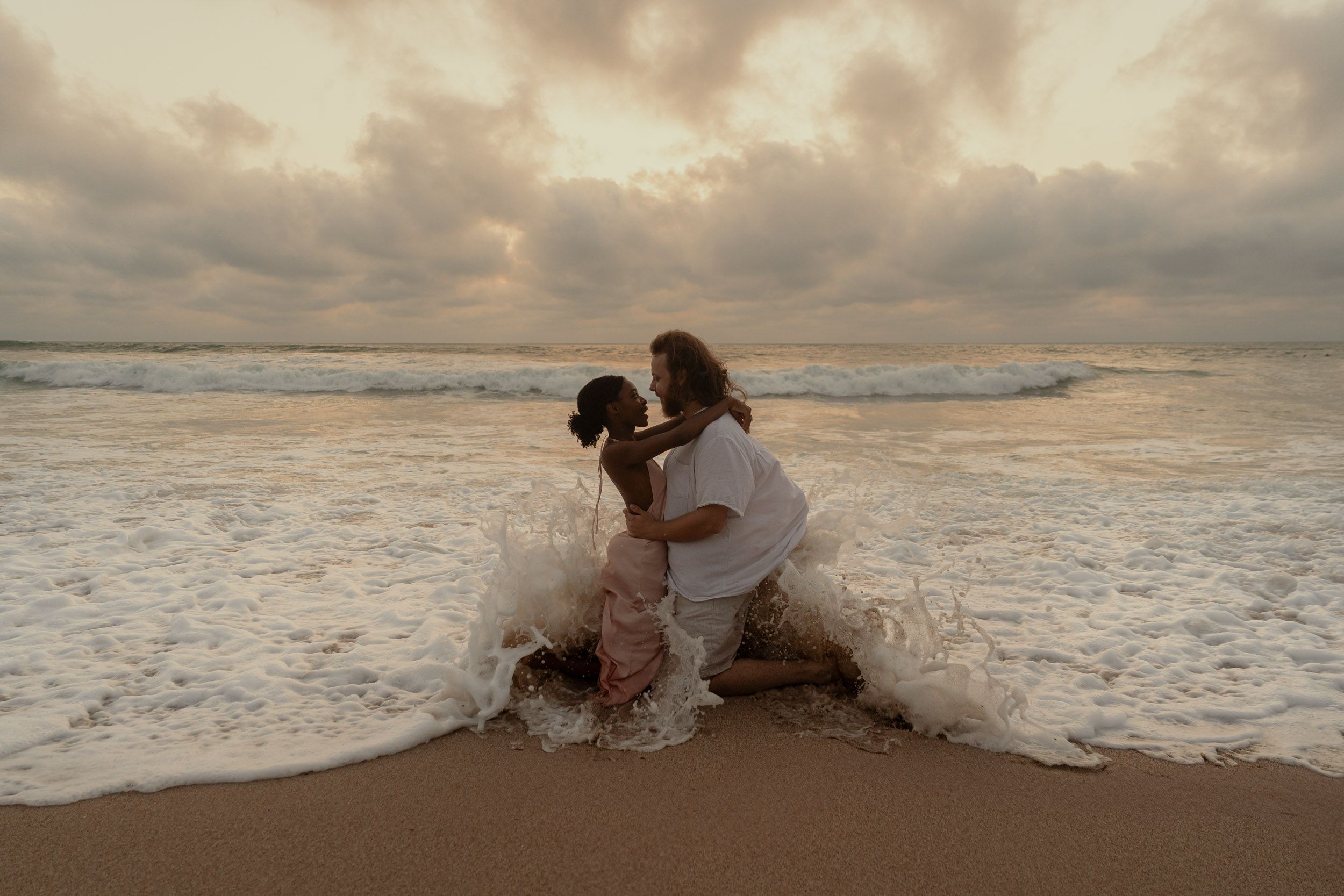 Couple playing in ocean waves at sunset on a sandy beach.