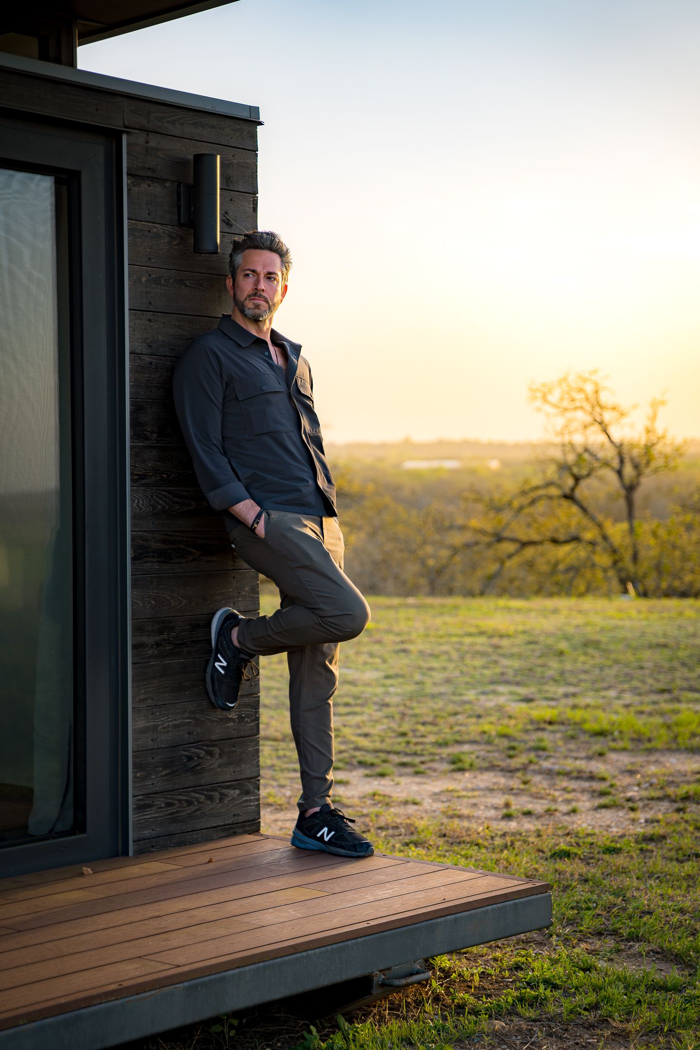 Person leaning against wooden cabin wall on deck at sunset with rural landscape in background.
