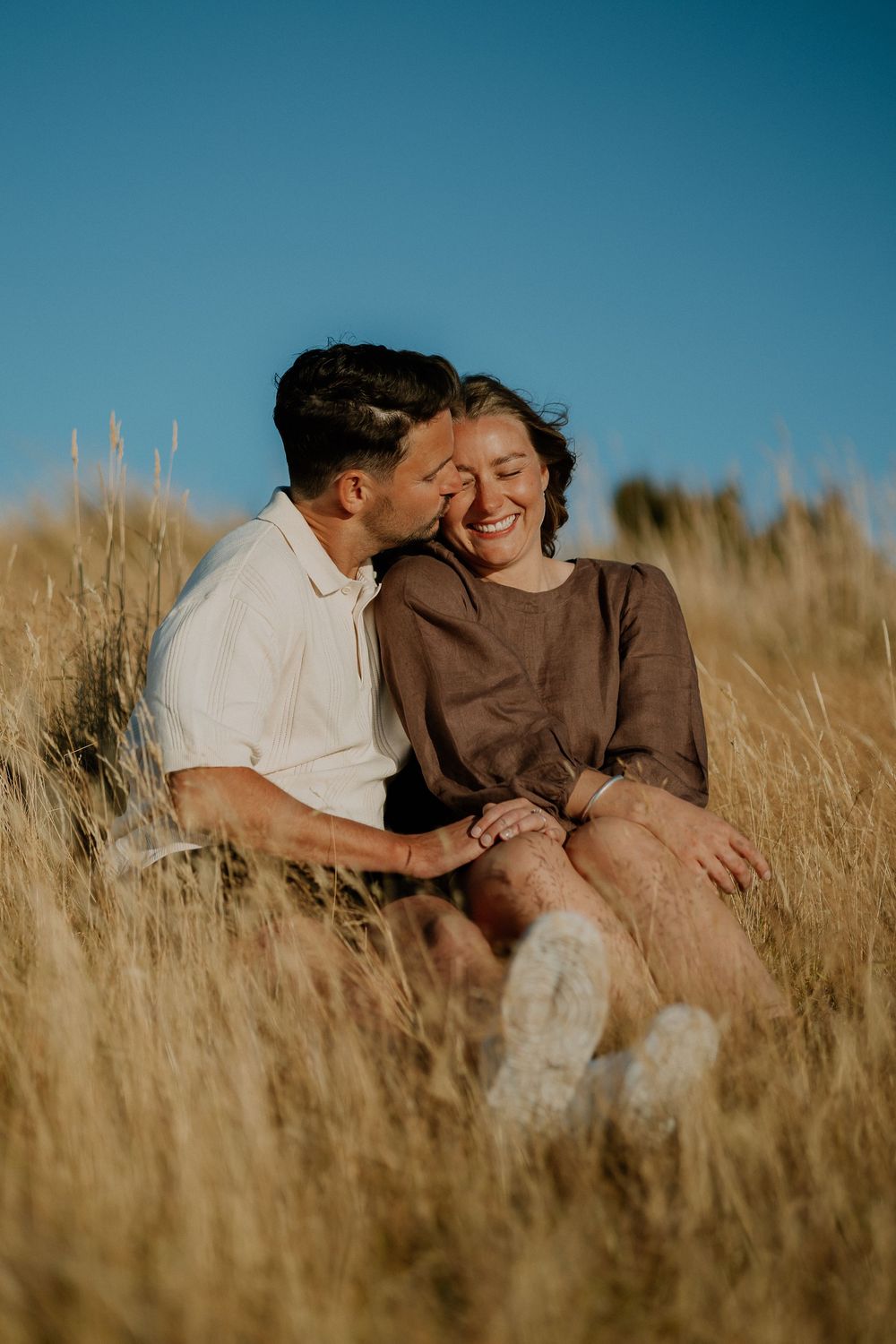 A couple shares an intimate moment while lying in tall golden grass during sunset.