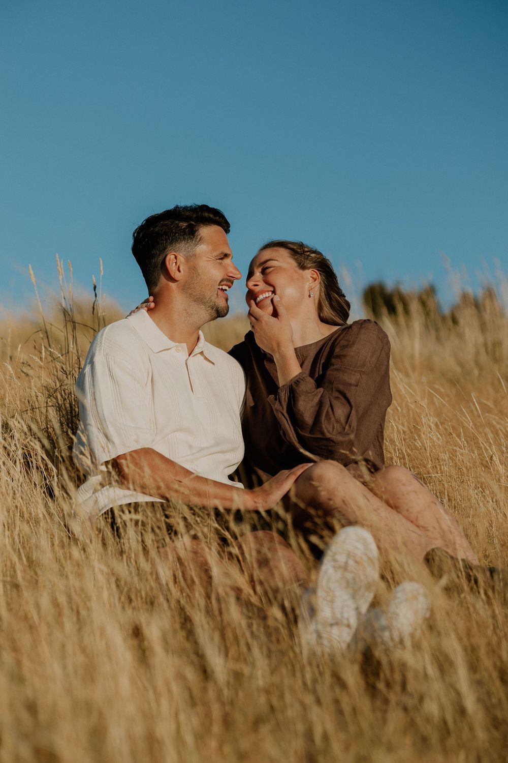 Romantic couple sharing intimate moments while sitting in tall grass against a blue sky.