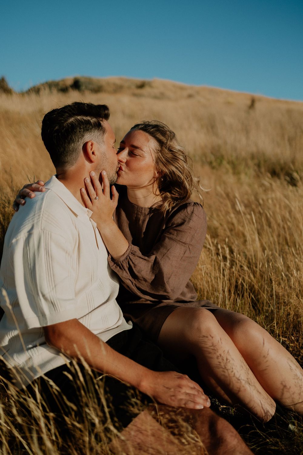 A romantic couple embraces while resting in a sunlit field of dry grass against a blue sky.