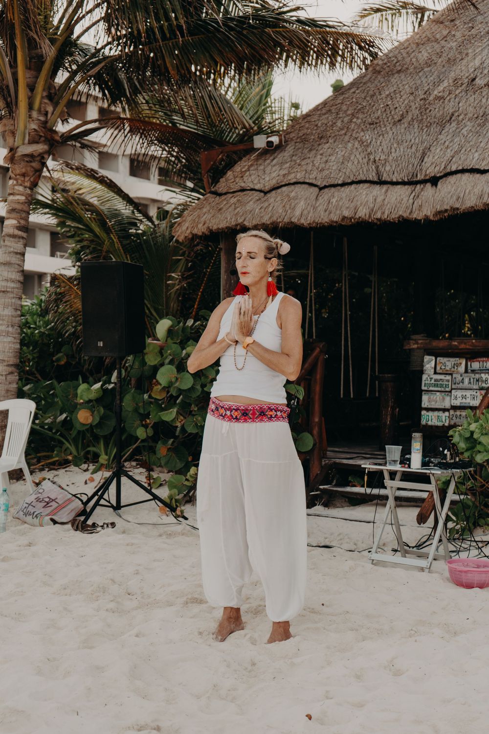 Person in white flowing outfit stands in prayer pose on sandy beach near thatched roof hut and palm trees.