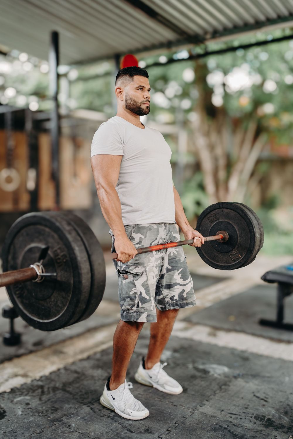 Person in red cap and camo shorts performs deadlift exercise with barbell in outdoor gym setting.