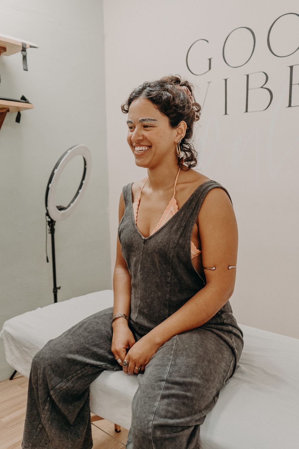 A person wearing a gray jumpsuit sits on a bed and laughs joyfully in a room with a ring light and 'GOOD VIBES' text.