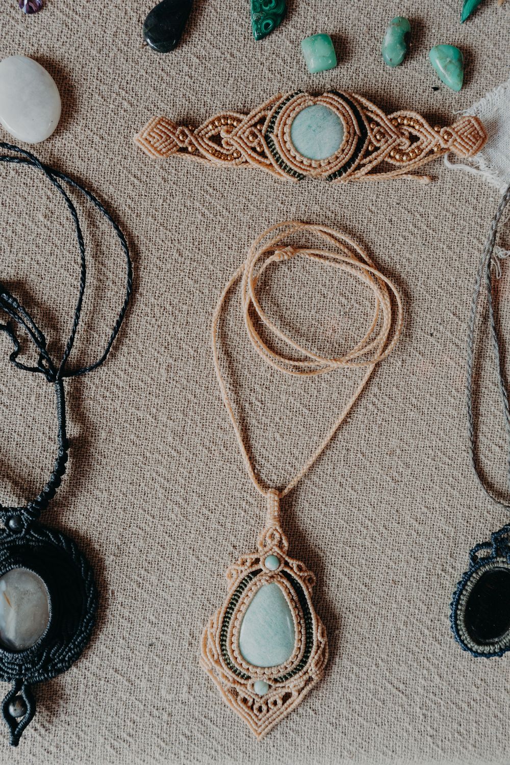 Handmade macrame jewelry featuring turquoise stones with beige cord necklaces and bracelets laid on textured fabric.