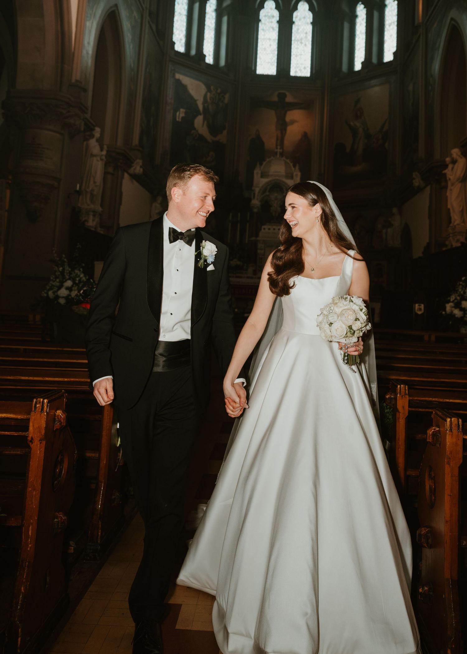 A couple walks down a grand cathedral aisle after their wedding ceremony in elegant formal attire.