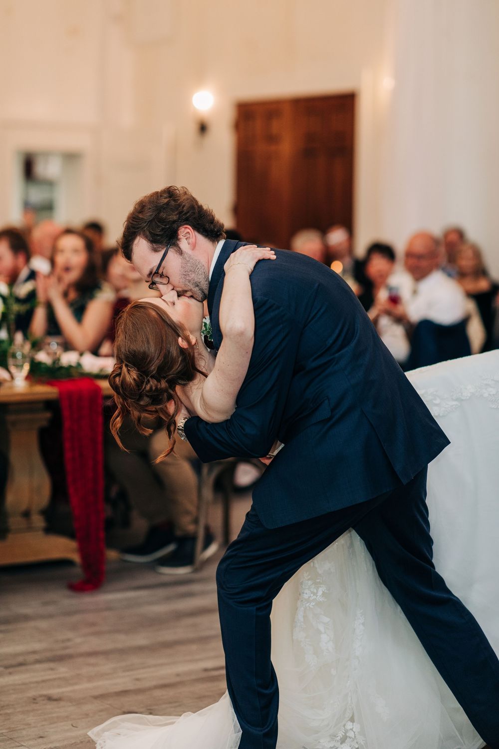 Couple performs dramatic dip during romantic first dance at wedding reception with guests watching in background.