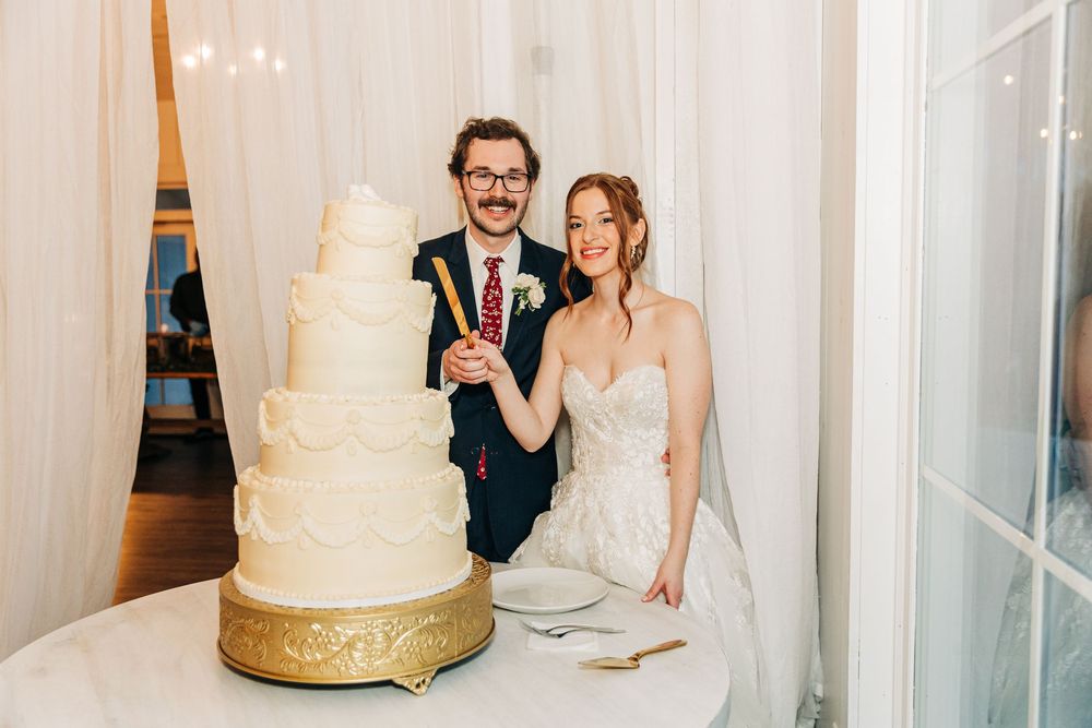 A couple stands together laughing while cutting their elegant white and gold tiered wedding cake at their reception.