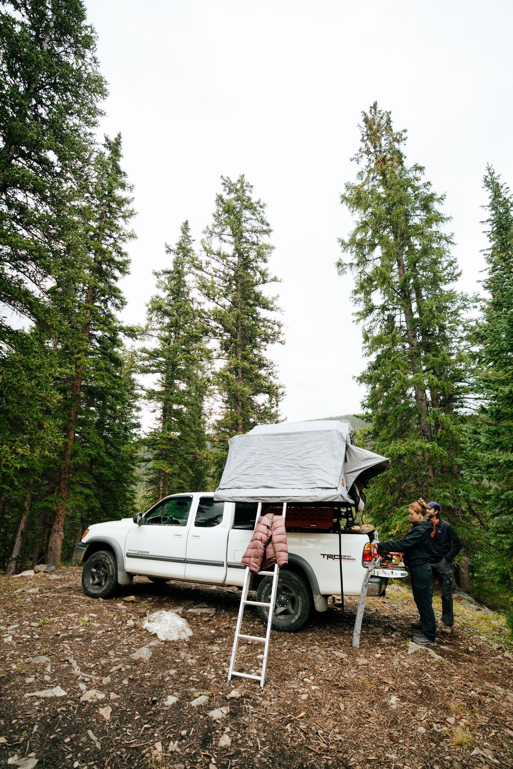 White pickup truck with rooftop tent parked in a forested mountain clearing with camping gear.