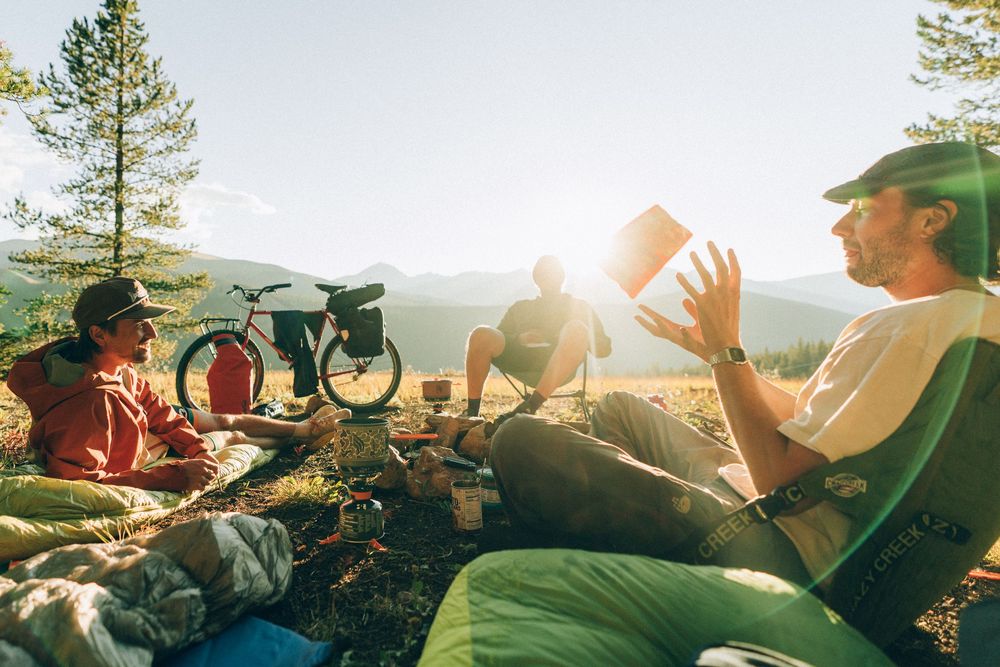 Group of outdoor enthusiasts enjoying conversation and laughter around a campsite in golden sunlight.