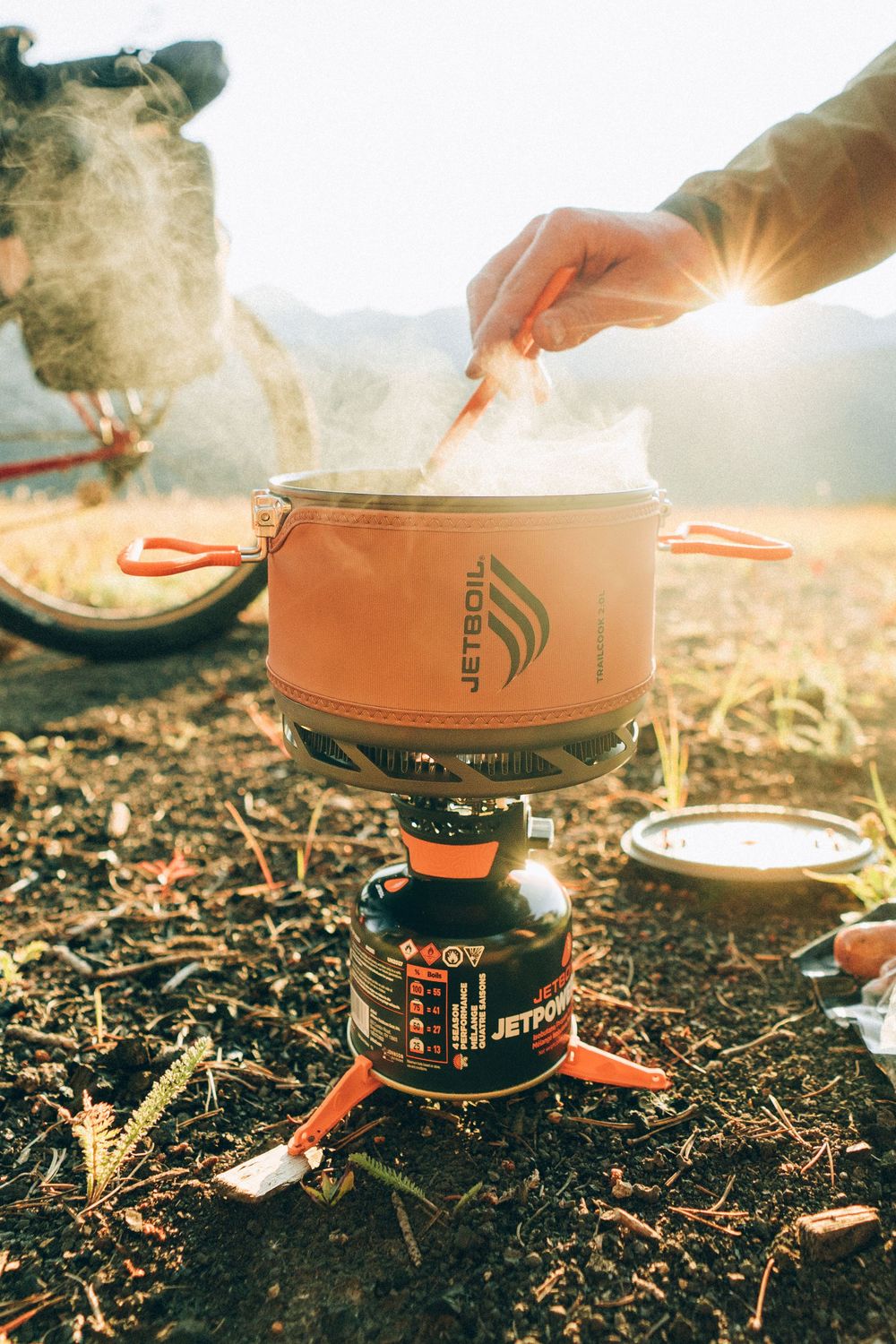 Camping stove burner being lit at sunset with hands adjusting flame controls in a natural outdoor setting.