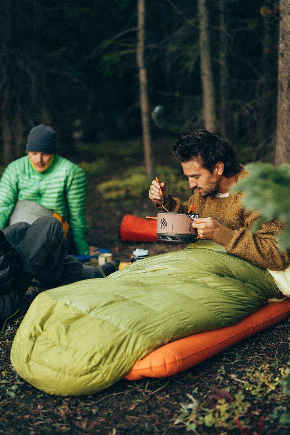 Camper relaxing in a lime green sleeping bag while drinking from a mug in a forest setting.
