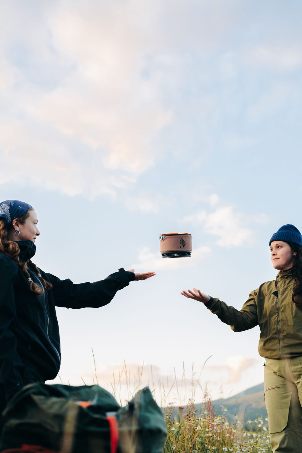 Two people in outdoor gear toss an object back and forth against a cloudy mountain backdrop.
