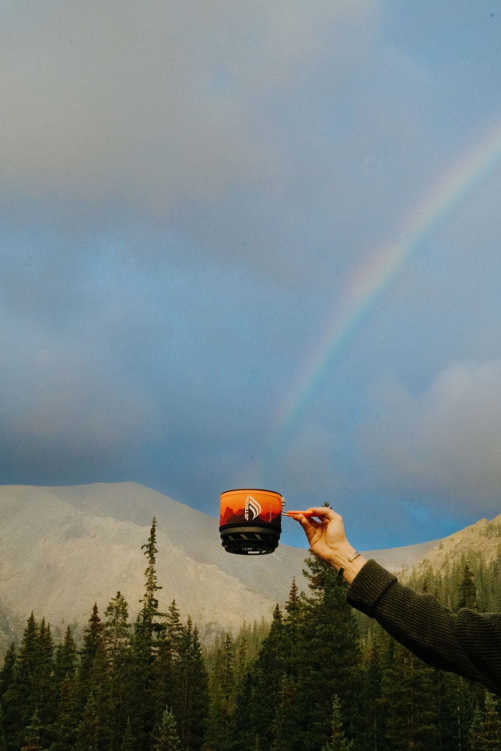 Rainbow arches over mountain peaks with camping cookware in the foreground.