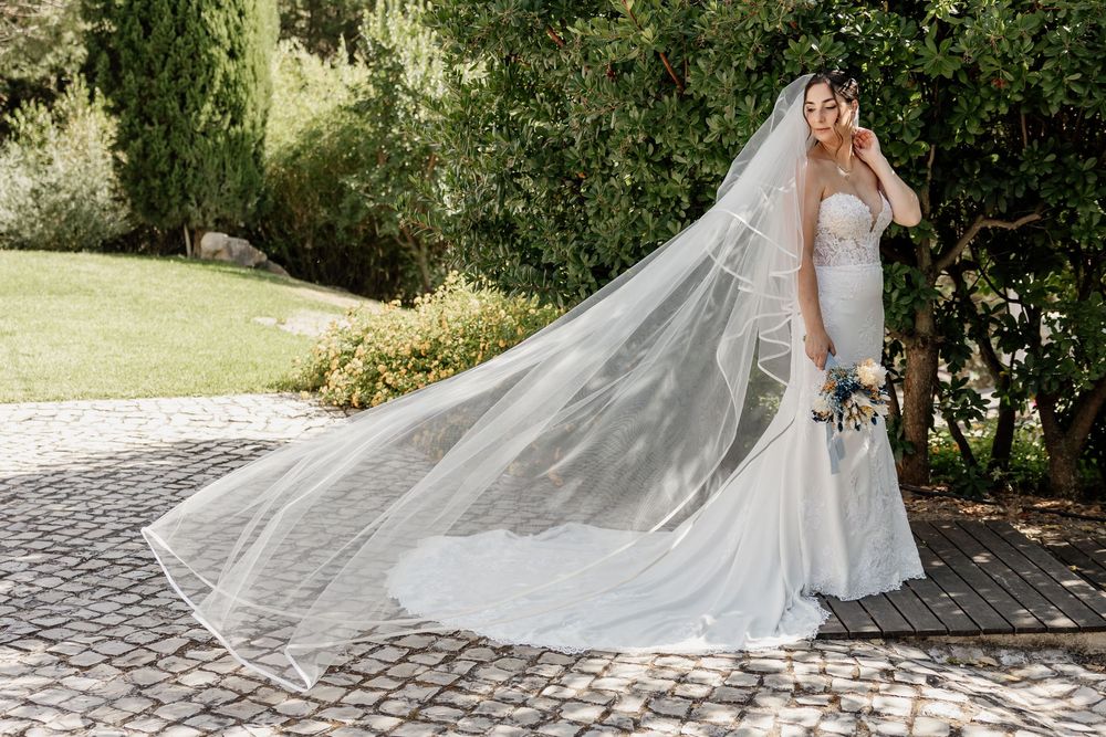 A flowing cathedral-length wedding veil trails behind a white bridal gown in a garden setting.