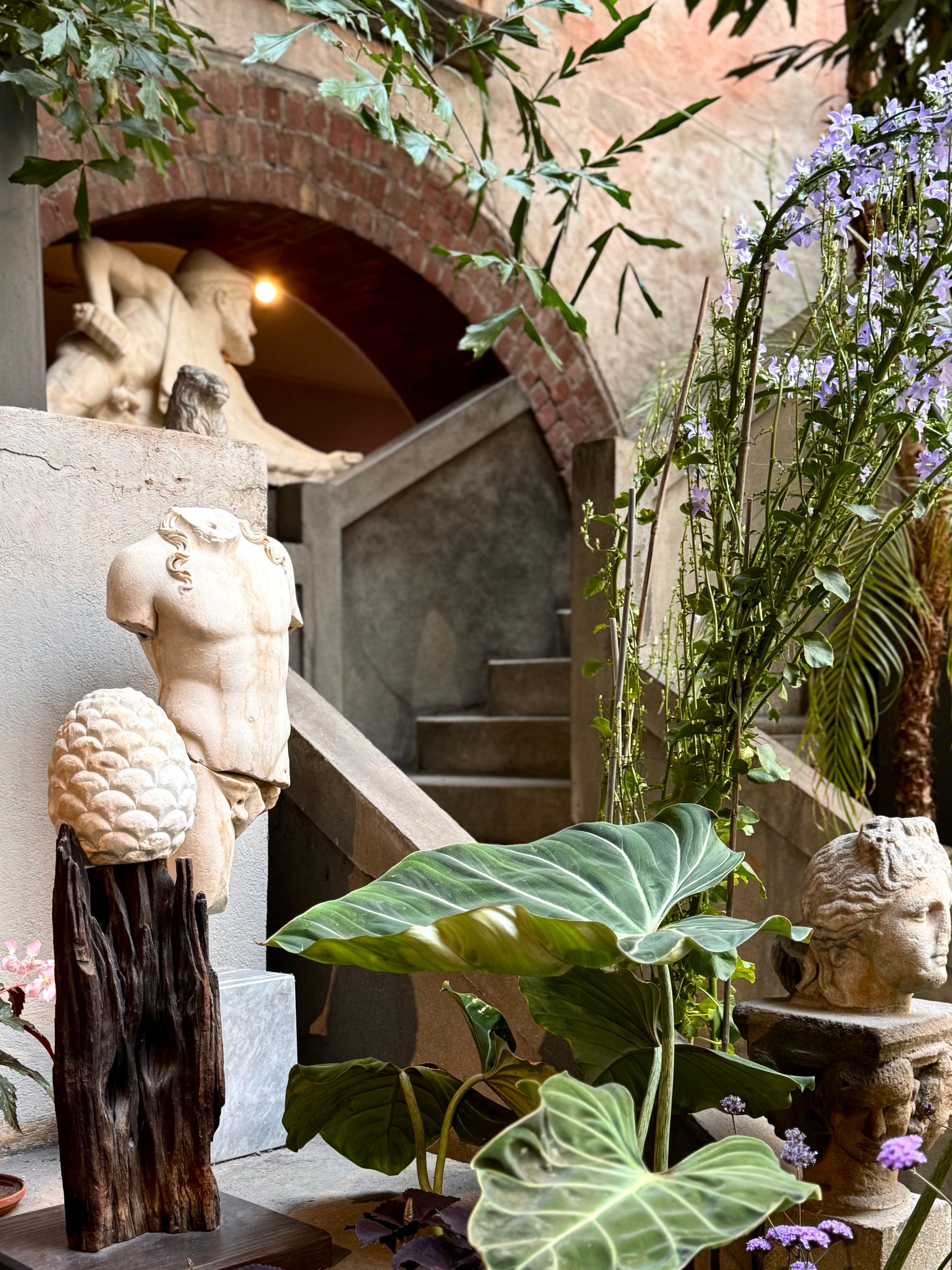 Classical stone archway with garden statuary, lush tropical plants and decorative elements in a Mediterranean courtyard setting.