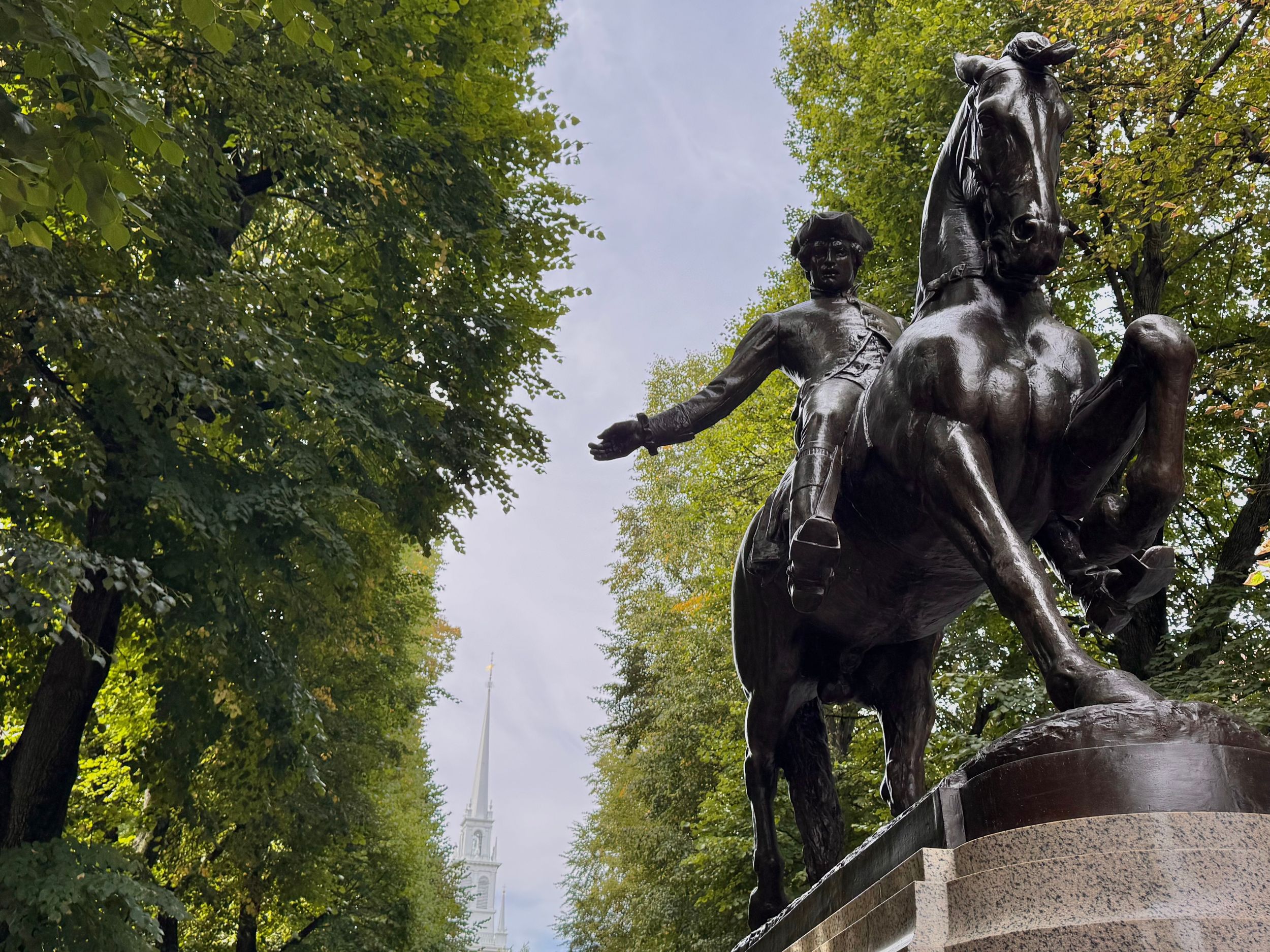 Bronze statue of a baseball player in pitching stance on stone pedestal surrounded by green trees in a park setting.
