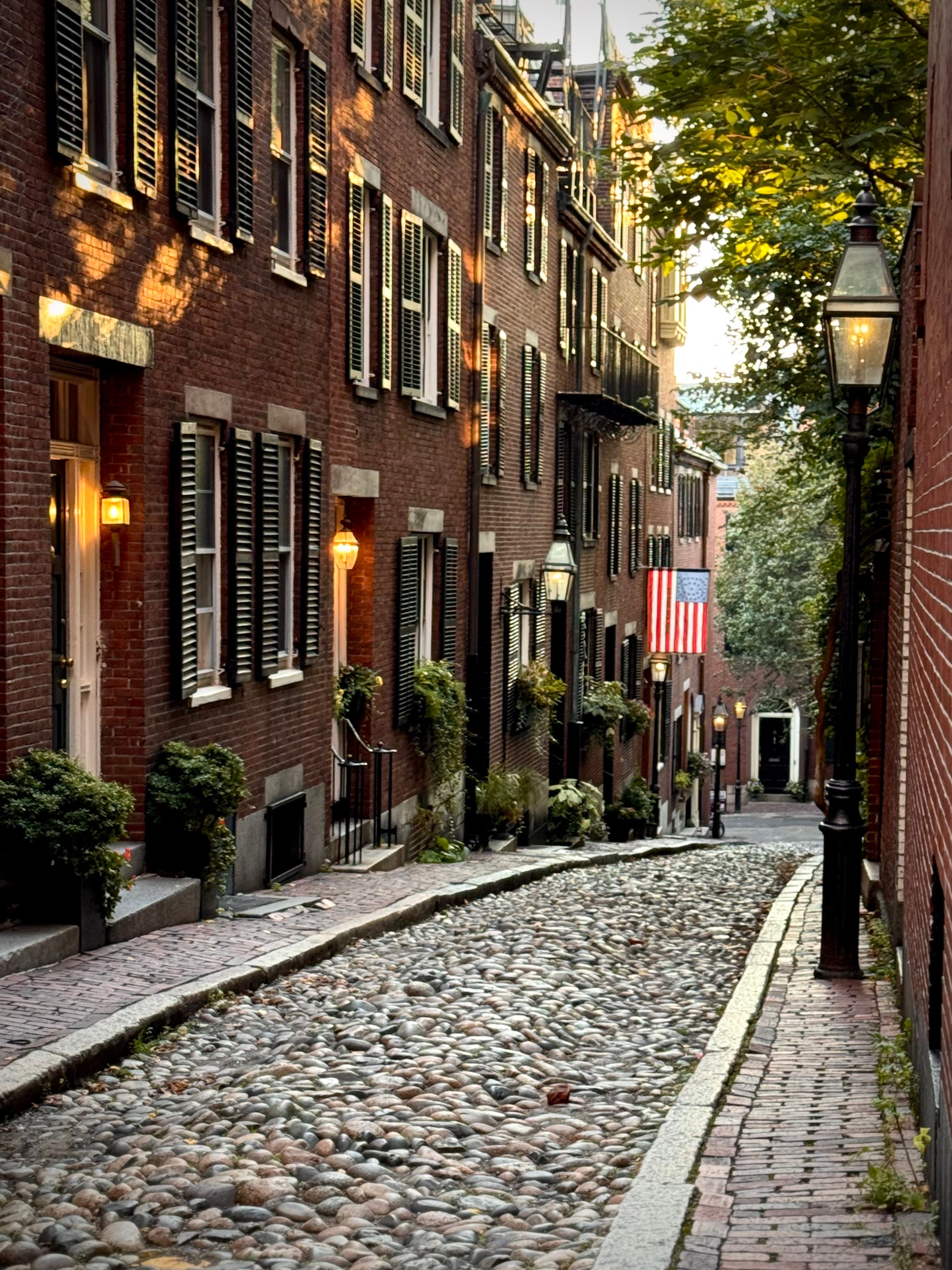Historic Acorn Street in Boston's Beacon Hill with cobblestones, red brick buildings, and an American flag at dusk.
