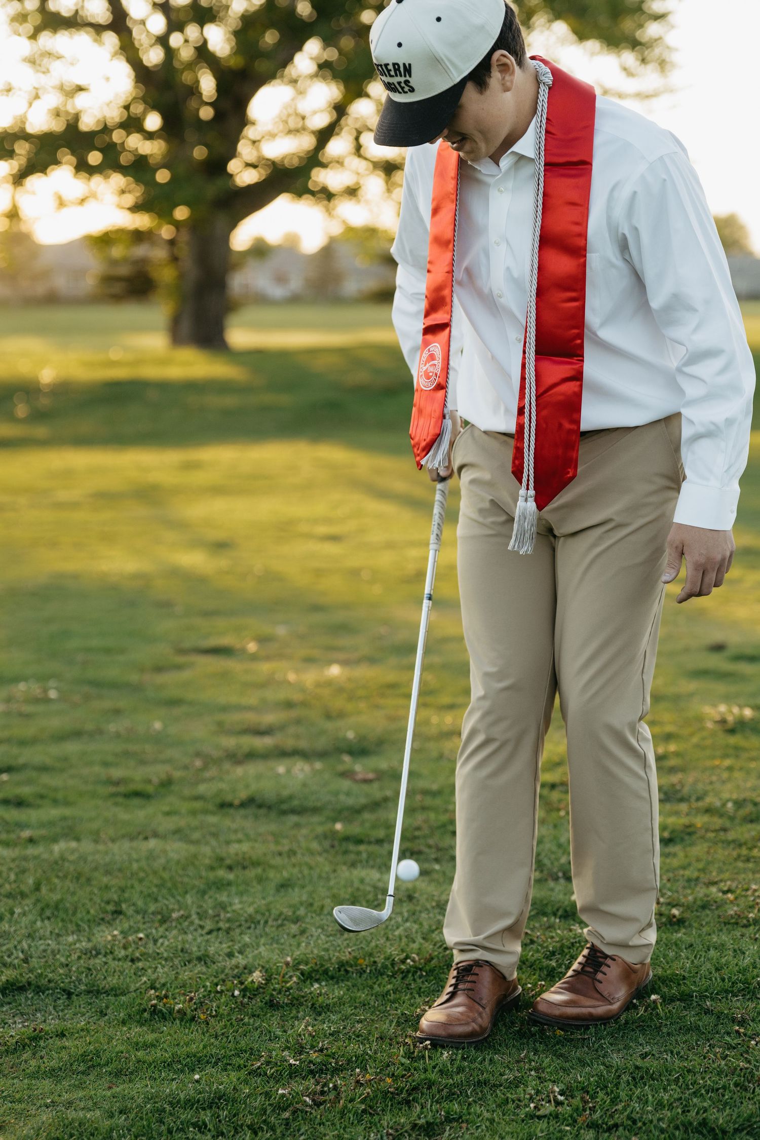 Young man in white shirt, khakis, and cap playing golf on a green course at sunset with a red sash.