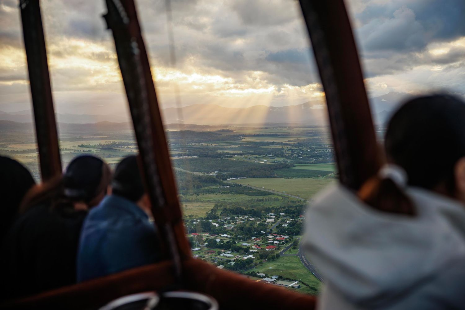 Aerial view from hot air balloon basket showing passengers gazing at sunlit green landscape below dramatic cloudy sky. Cairns Australia