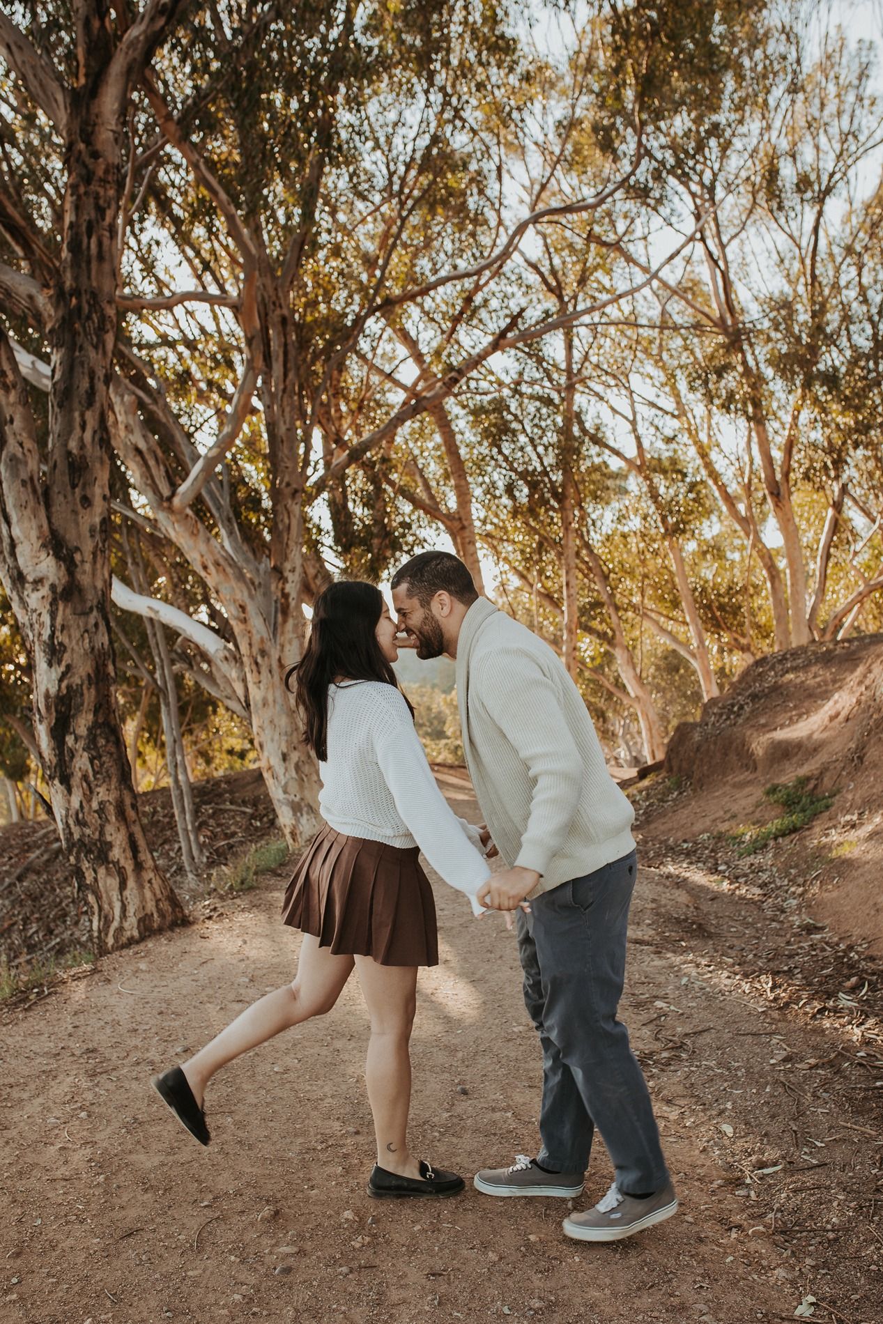 A couple shares a romantic moment on a dirt path surrounded by eucalyptus trees in warm autumn light at Will Rogers State Park.