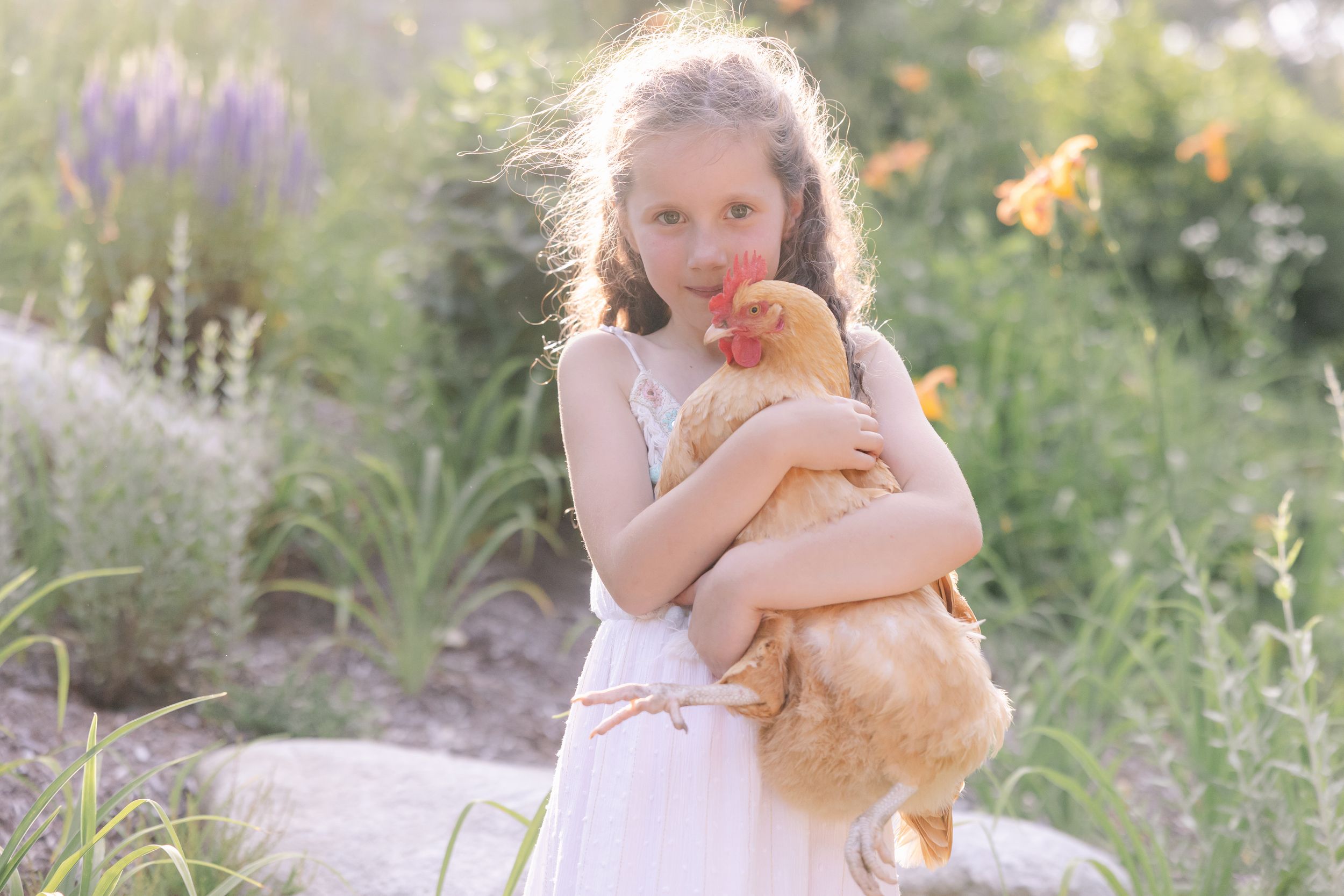 A person in a flowing white dress holds a golden retriever puppy in a dreamy outdoor garden setting with purple flowers.