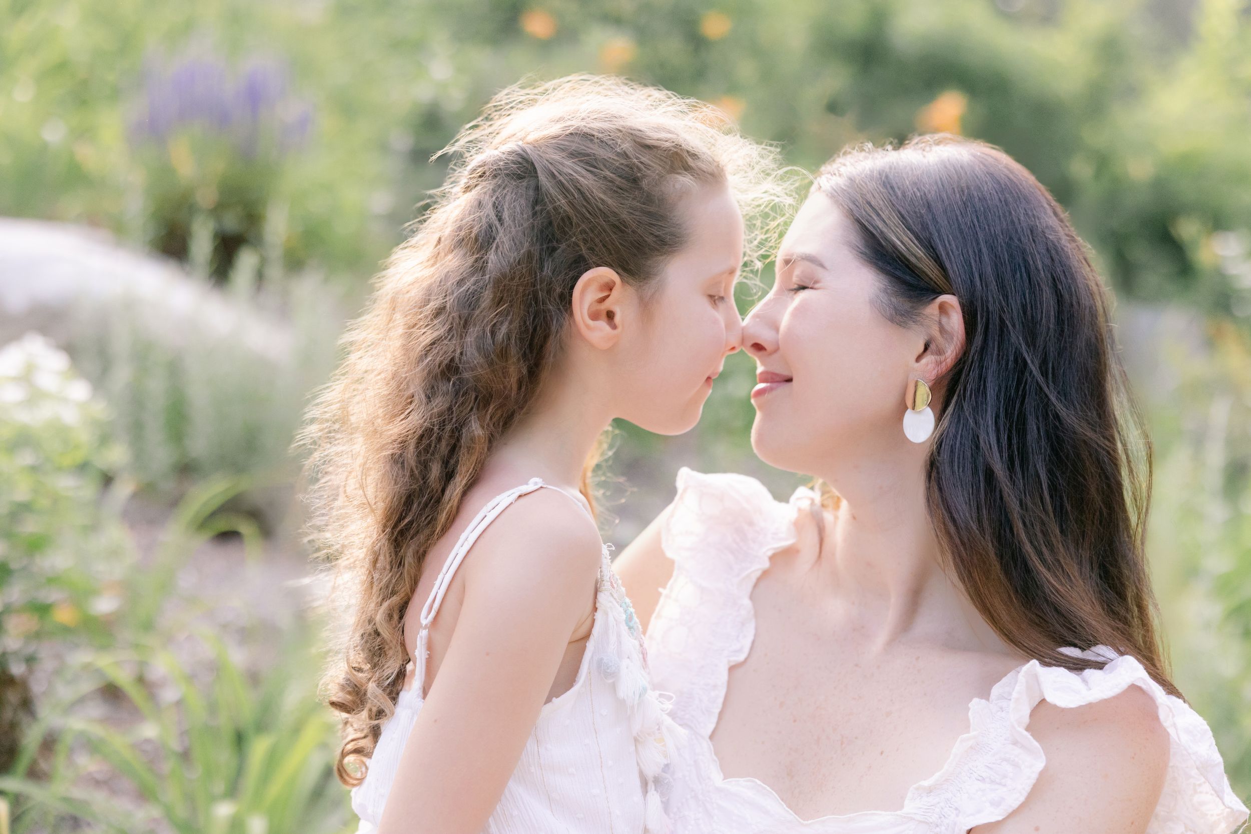 Two people in white lace dresses share a joyful moment together in a sunlit garden with flowers.