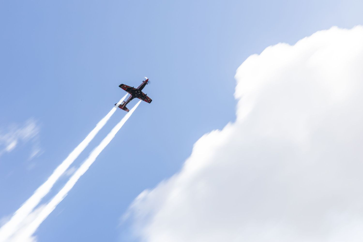 A small airplane flies through a bright blue sky leaving white contrails behind it.