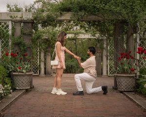 Man kneeling on one knee holding woman's hand during a romantic garden proposal with red tulips.