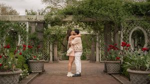 Happy couple embracing and kissing under a white garden arbor surrounded by lush greenery and red tulips.