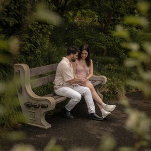 Couple sitting closely together on a park bench surrounded by dense green trees and foliage.
