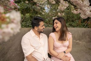 Smiling South Asian couple sitting on a bench laughing together beneath pink cherry blossom trees.