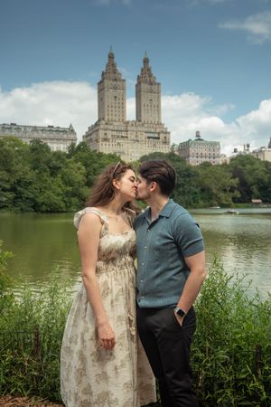 Couple sharing a kiss by a lake in Central Park with iconic NYC skyline buildings in background.