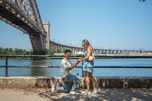 Man on one knee proposing to his girlfriend by the waterfront with a large bridge in the background.