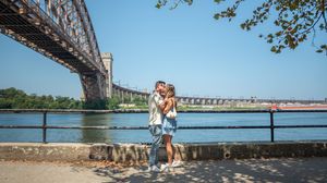 Couple kissing romantically by the waterfront with a large steel arch bridge and blue sky behind them.