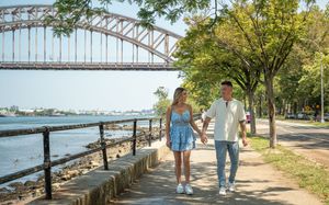 Happy couple walking hand in hand along a waterfront path with a steel arch bridge in the background.