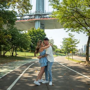Couple embracing and kissing on a path beneath a large bridge surrounded by green trees in a park.