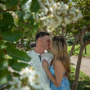 Couple sharing a kiss framed by white flowering tree branches in a lush green park setting.