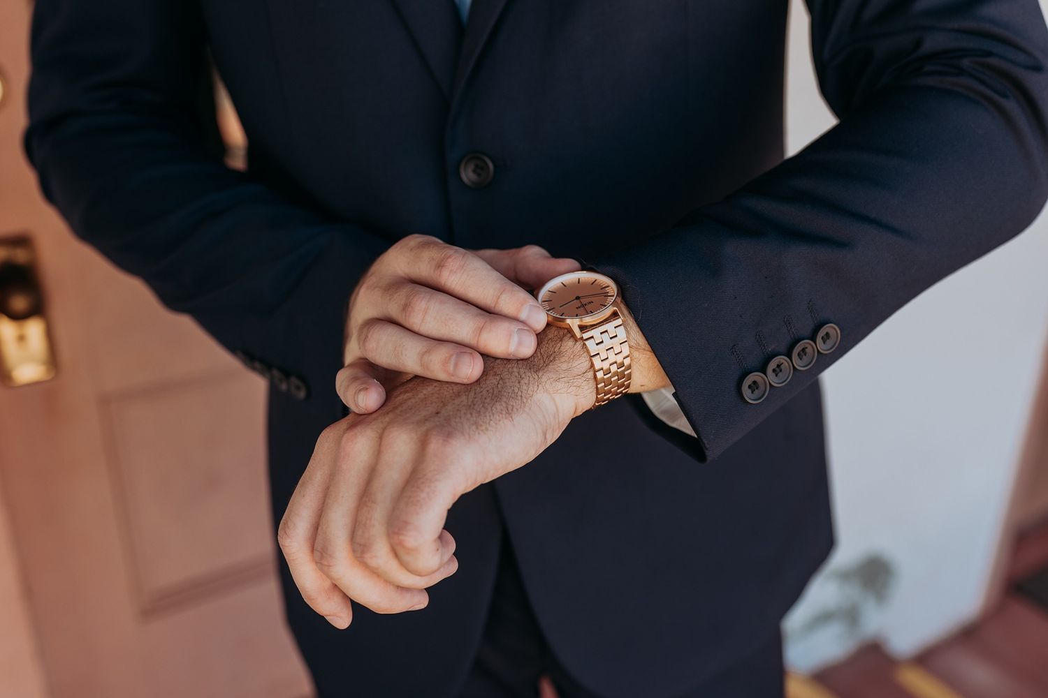 Groom in navy suit checking rose gold watch on wrist, elegant formal attire for his wedding.