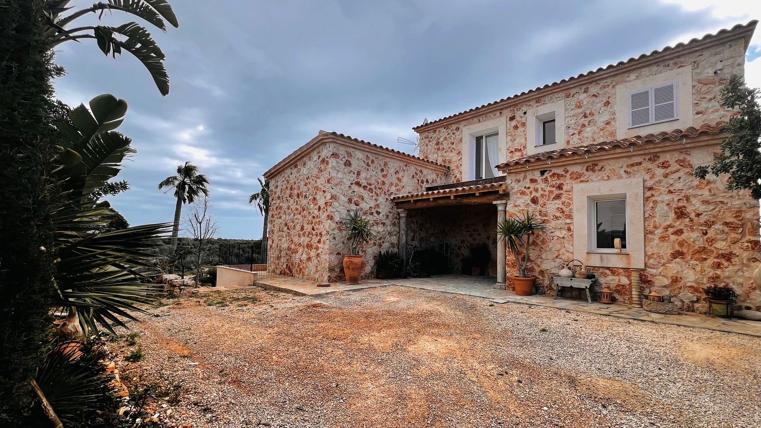 A rustic stone building with a terracotta roof, surrounded by lush vegetation and a dirt driveway.