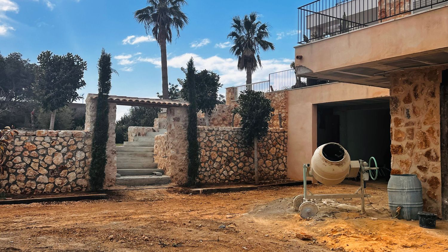 A stone wall and archway lead to a courtyard with palm trees and a large ceramic pot in the foreground.