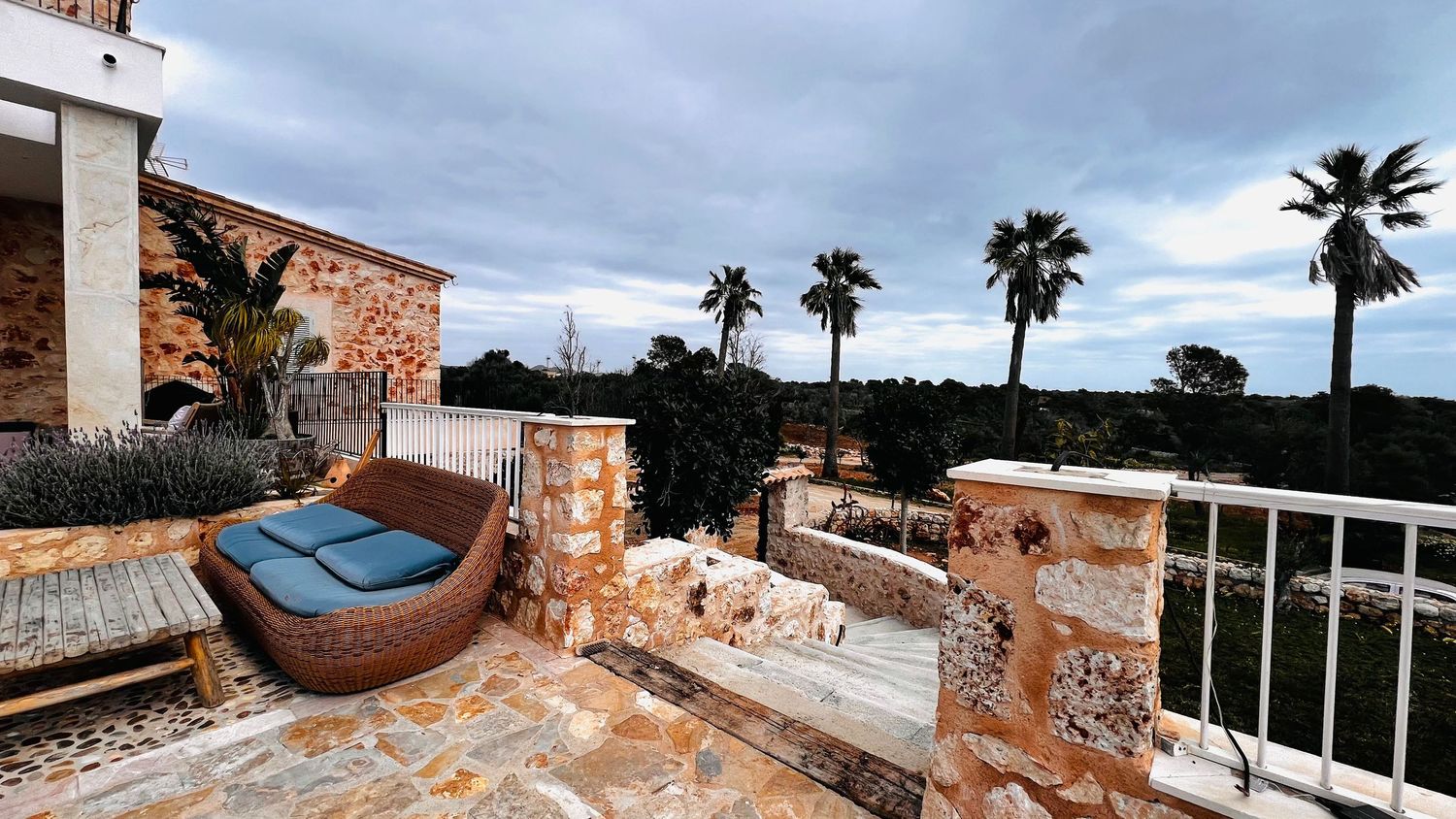 A patio area with outdoor furniture and a stone wall, with palm trees and a cloudy sky in the background.