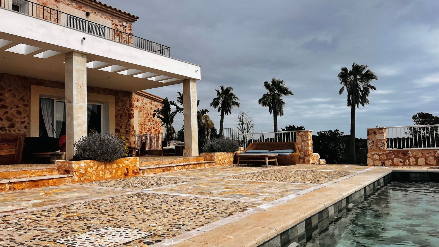 A scenic outdoor pool area with palm trees, surrounded by a stone building and paved walkways.