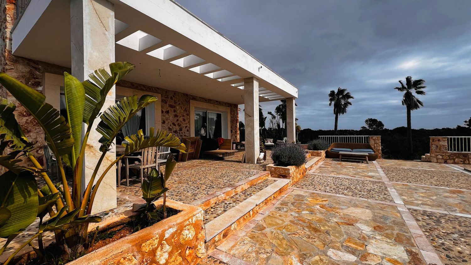 A covered patio or porch with stone walls and a tiled roof, surrounded by palm trees and other tropical plants.