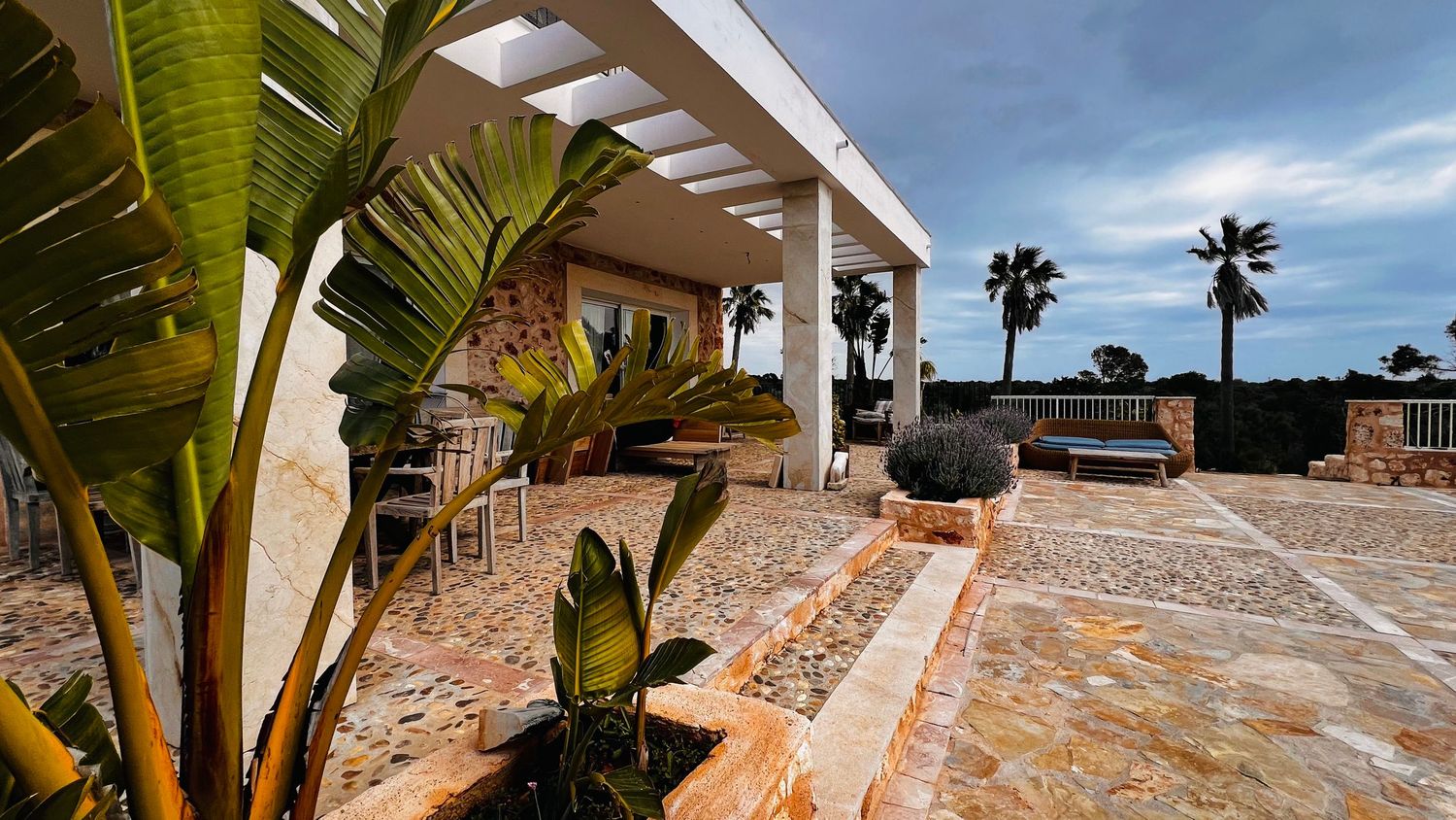 Outdoor patio with palm trees, tiled floor, and covered seating area with a scenic view.