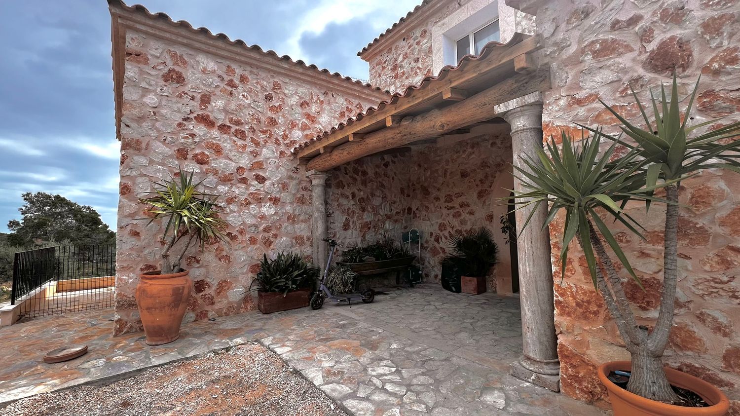 A rustic stone building with a tiled roof, surrounded by potted plants and palm trees in a Mediterranean-style setting.