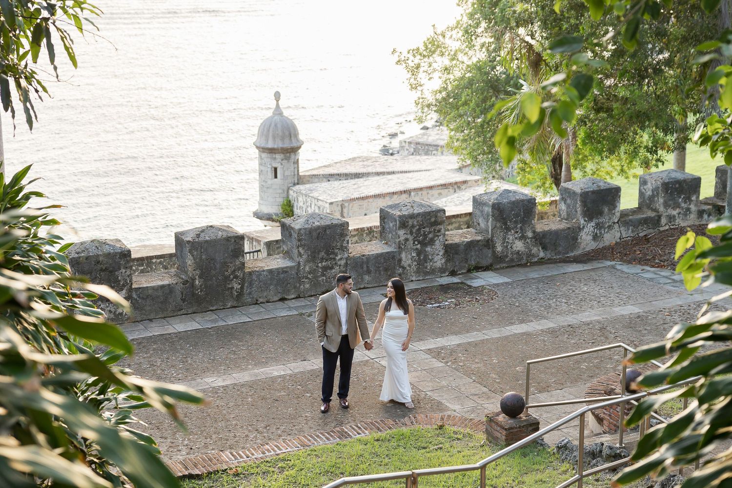 A couple dressed in formal attire stand in a garden setting within a fort in Old San Juan and greenery.