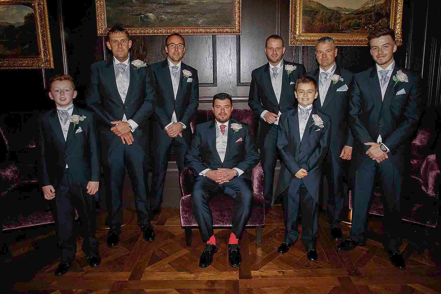 A wedding party in navy suits poses in a formal setting with wood floors and framed artwork on the walls.
