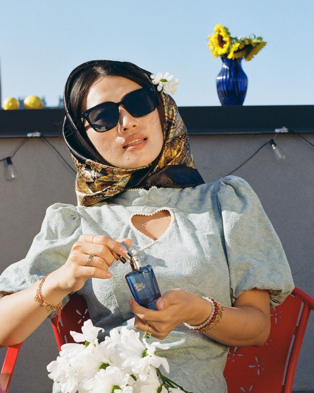Stylish woman wearing sunglasses and floral headscarf holding a blue perfume bottle while seated outdoors.