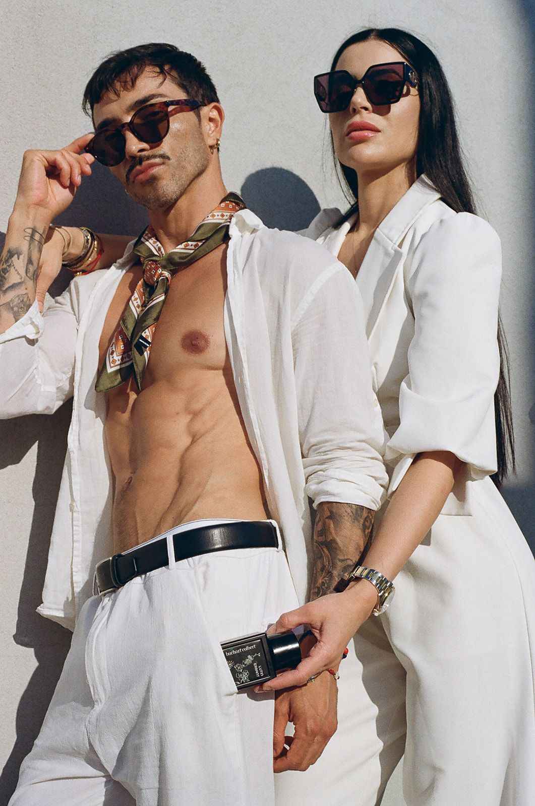 Stylish couple in white outfits and sunglasses posing outdoors, man with open shirt and scarf, woman holding a clutch.